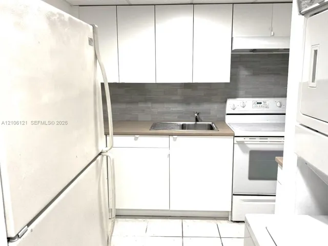 White cabinetry, neutral-toned backsplash, built-in range with overhead vent, stainless steel sink, and tile flooring