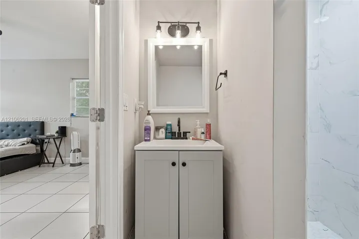 Light grey Shaker-style vanity with a rectangular sink, matte black faucet, framed mirror, and three-bulb vanity light fixture