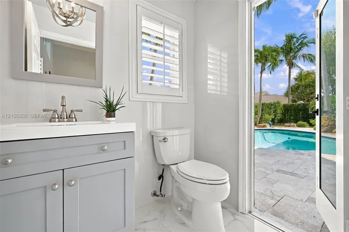 Bathroom featuring a gray vanity with a white countertop, wall-mounted mirror, white plantation shutters, and glass-paneled door opening to a stone-paved patio with an in-ground pool