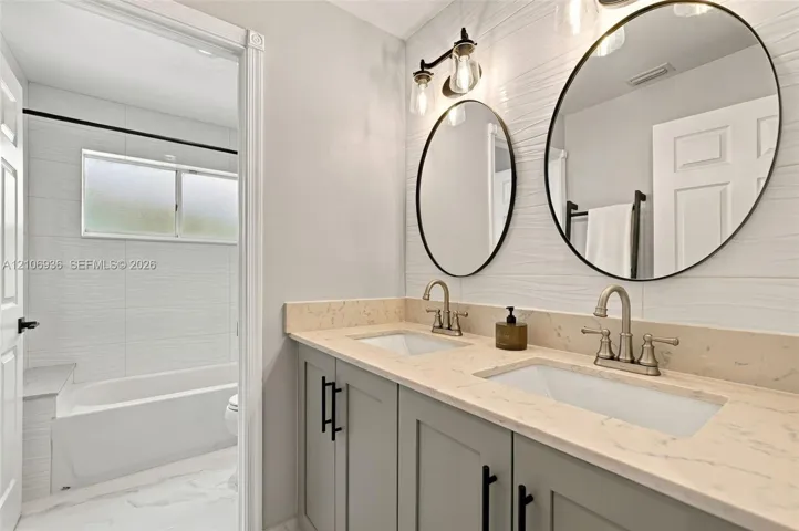 Dual-sink vanity with light-toned stone countertop, shaker-style cabinetry, matte black hardware, oval mirrors, and white textured tile walls