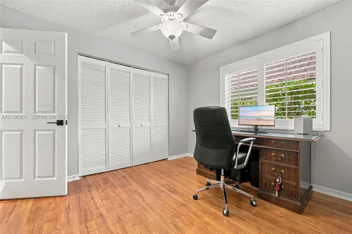 Wood-finish flooring, white louvered closet doors, multi-panel interior door with matte black hardware, dual-pane window featuring white plantation shutters, and ceiling fan with integrated light fixture