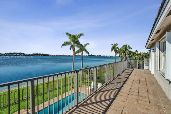 Elevated terrace with tile flooring and metal railings, overlooking a rectangular swimming pool, manicured lawn, and waterfront expanse