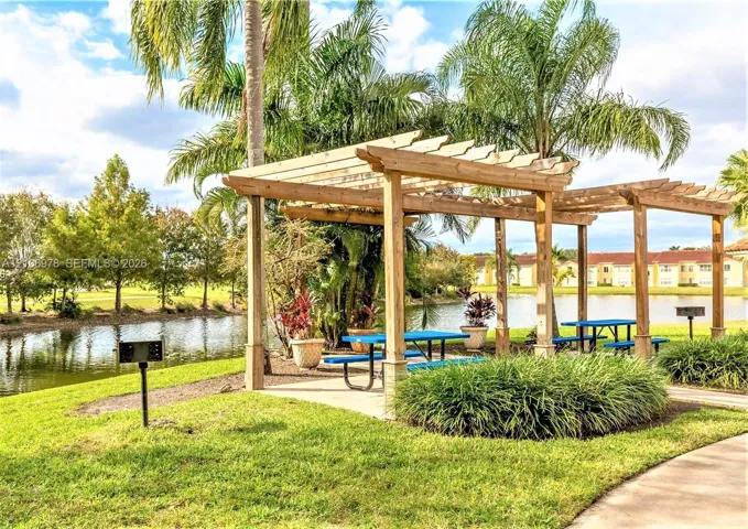 Outdoor common area featuring wood-frame pergolas, concrete patio surfaces, and charcoal grills, positioned alongside a lake with mature landscaping