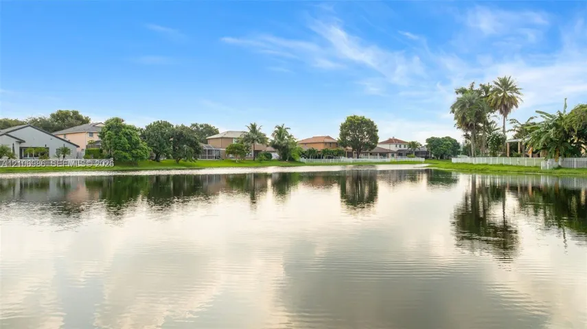 Waterfront setting featuring a calm pond, mature palm trees, perimeter white fencing, and expansive green space