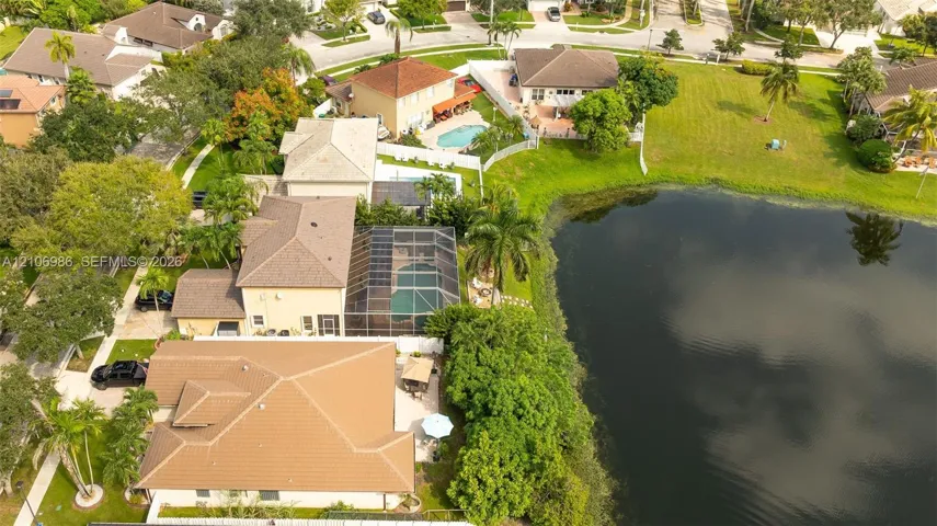 Aerial perspective featuring a waterfront property with a screened-in pool enclosure, concrete tile roofing, paved driveway, and mature landscaping