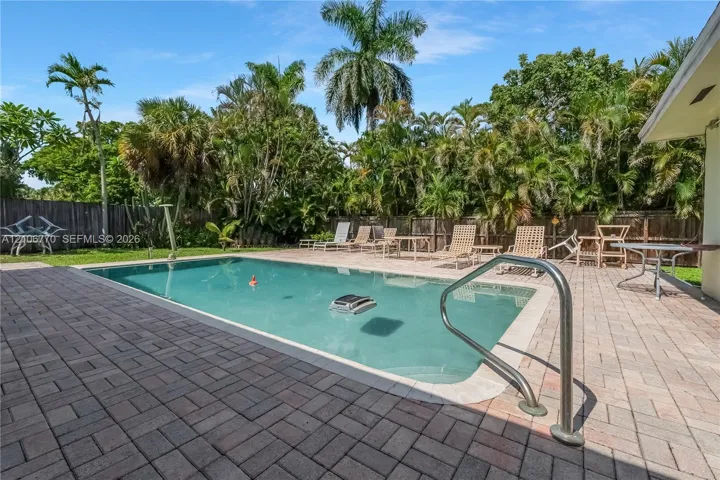 In-ground swimming pool with a metal handrail, surrounded by a paved stone patio, enclosed by a wood privacy fence and mature palm trees