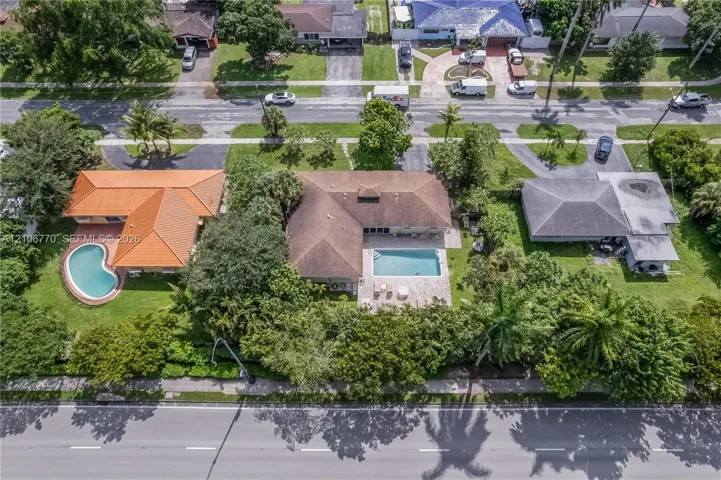 Aerial perspective featuring a shingled roofline, rectangular in-ground swimming pool, paved patio, and established perimeter landscaping on a lot with mature trees
