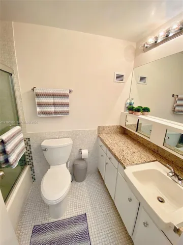 Bathroom featuring a vanity with stone-finish countertop, integrated sink, large wall-mounted mirror, mosaic tile surround, and glass-enclosed shower