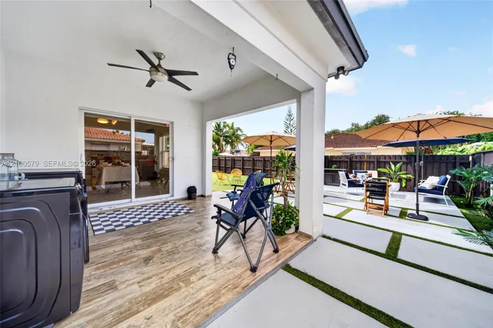 Covered patio featuring wood-finish flooring, a ceiling fan, and sliding glass door access, complemented by an expansive concrete paver patio with turf inlays and a perimeter privacy fence
