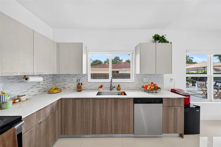 Kitchen featuring two-tone flat-panel cabinetry, light-toned stone-finish countertops, multi-textured glass tile backsplash, stainless steel dishwasher, and a dual-pane window