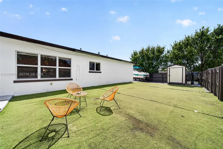 Backyard featuring synthetic turf, white stucco exterior, multipane windows, and a detached storage shed enclosed by wood fencing