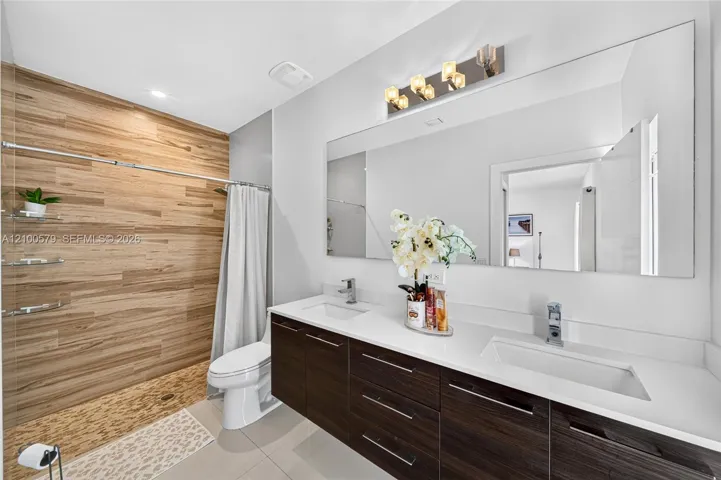 Dual-sink vanity with a solid-surface countertop, wood-finish cabinetry, full-width wall mirror, and shower area featuring wood-finish wall tile and pebble-texture flooring