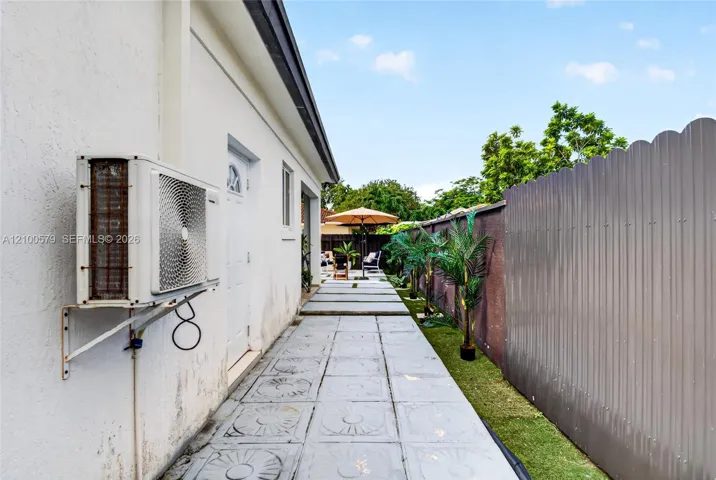 Side exterior featuring concrete paver flooring, white stucco wall finish, and a corrugated metal privacy fence