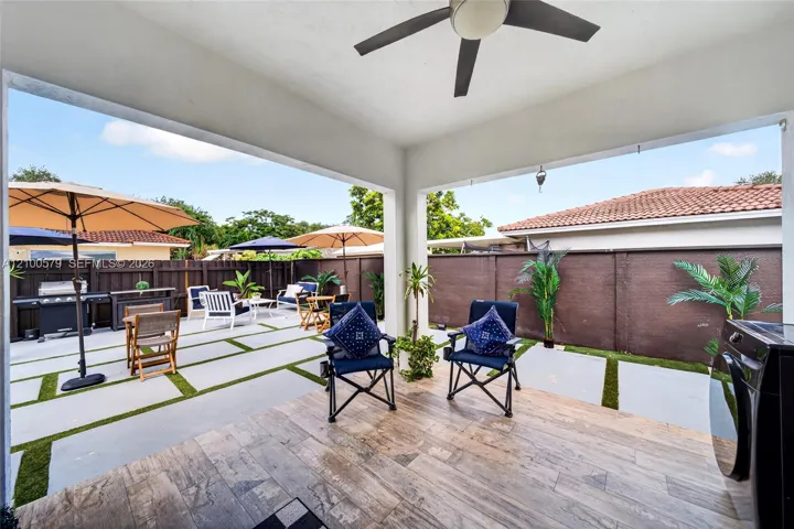 Covered patio featuring wood-finish flooring and a ceiling fan, opening to a concrete paver hardscape with integrated turf accents and dark wood perimeter fencing