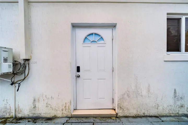 White exterior wall with textured stucco finish, featuring a four-panel entry door with a semi-circular transom window and a rectangular window with a white frame