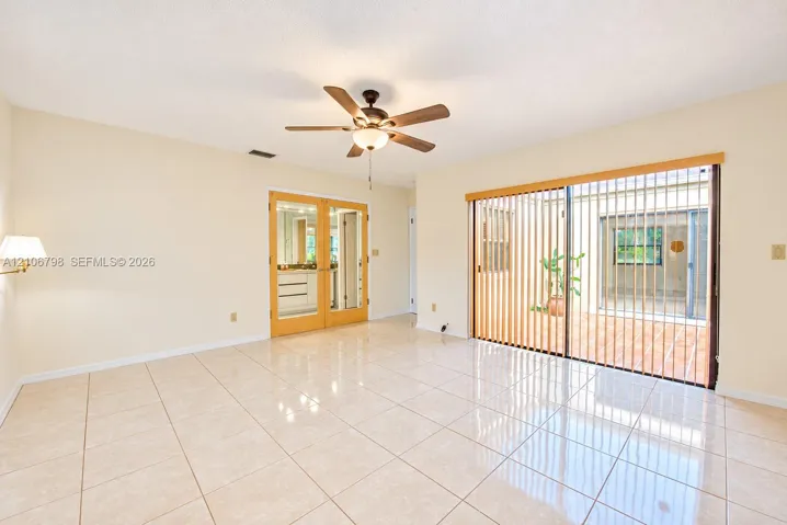 Spacious room featuring polished tile flooring, wood-framed French doors, and large sliding glass doors opening to an enclosed patio area, complemented by a central ceiling fan