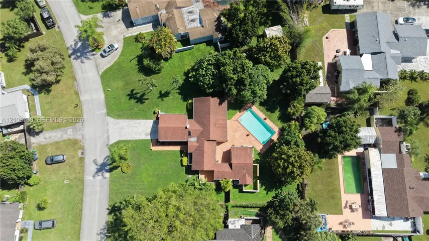 Aerial perspective of a property featuring a multi-gabled shingle roof, paved driveway, expansive lawn, mature landscaping, and an inground swimming pool with a paved patio