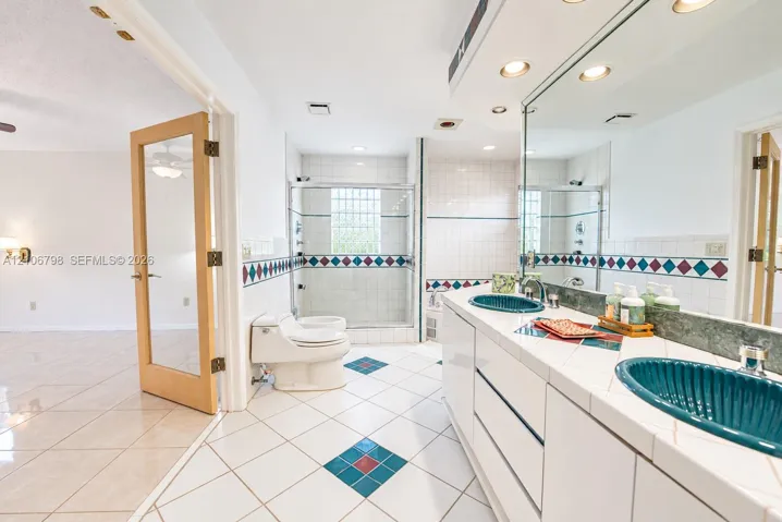Bathroom featuring a dual-sink vanity with tiled countertops, two ribbed teal vessel sinks, a glass-enclosed shower with glass block window, and white tile flooring with decorative accent insets