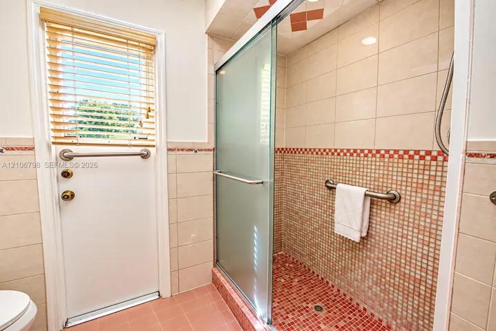 Walk-in shower featuring a glass sliding door, mosaic tile detailing, wall-mounted safety grab bars, and neutral wall tile