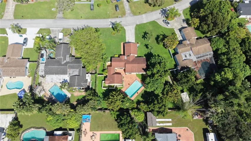 Aerial view featuring a multi-gabled residence with a terracotta-toned shingle roof, a rectangular inground swimming pool, paved patio area, and mature landscaping on a corner lot