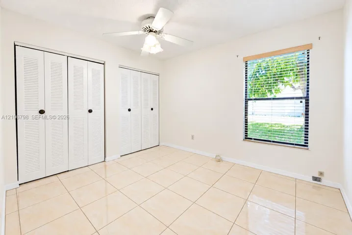 Interior room featuring large-format neutral tile flooring, dual louvered-style closets, a white ceiling fan with integrated light fixture, and a single window with wood-finish trim