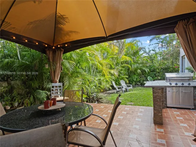 Outdoor patio featuring terracotta-style tile flooring, built-in outdoor kitchen with stainless steel grill, stone-finish base, and landscaped backyard with mature greenery