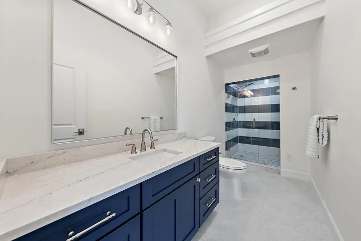 Navy blue vanity with light-toned stone countertop, integrated sink, expansive wall-mounted mirror, and walk-in shower with horizontal tile pattern