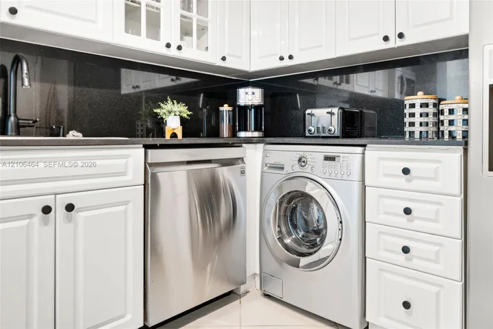 White cabinetry with raised-panel doors, solid-surface dark countertops, full-height dark backsplash, stainless steel dishwasher, and integrated front-loading washer