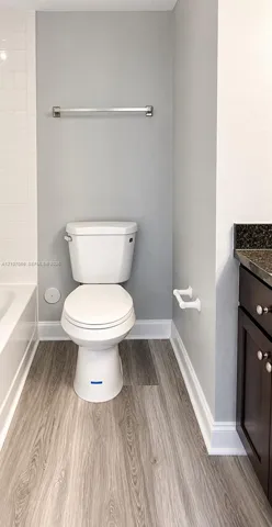 Bathroom featuring wood-finish flooring, a dark-toned vanity with stone-surfaced countertop, a white porcelain toilet, and white wall tile surround
