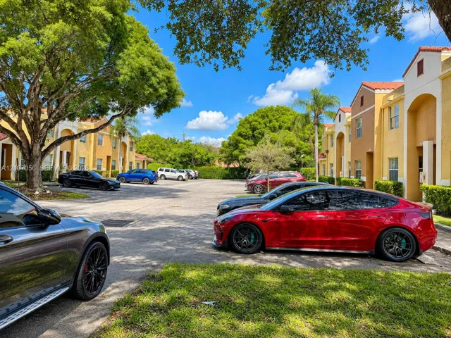 Asphalt surface parking lot featuring mature trees and landscaped shrubbery set against multi-story exterior architecture with arched entryways and tile roofing