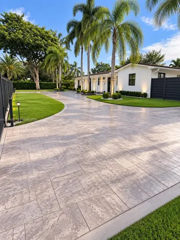 Paved motor court featuring large-format stone-finish pavers, manicured lawn areas, mature palm trees, and perimeter fencing
