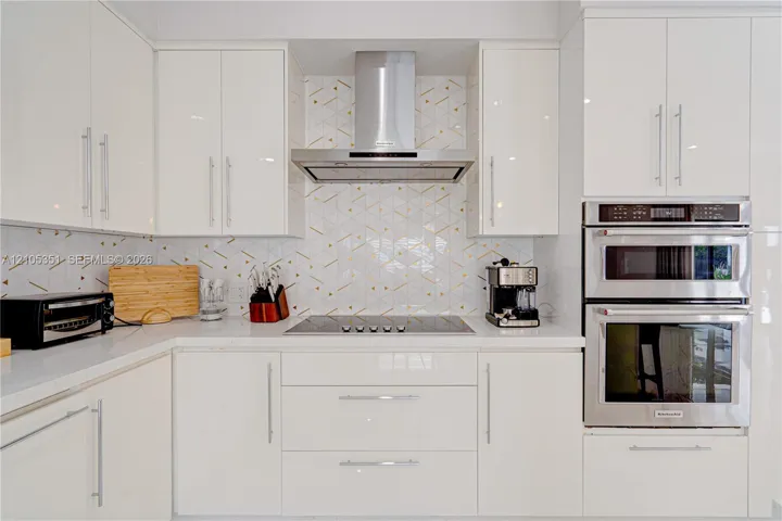Kitchen featuring high-gloss white cabinetry, geometric patterned backsplash, stainless steel double wall oven, stainless steel range hood, and solid-surface countertops