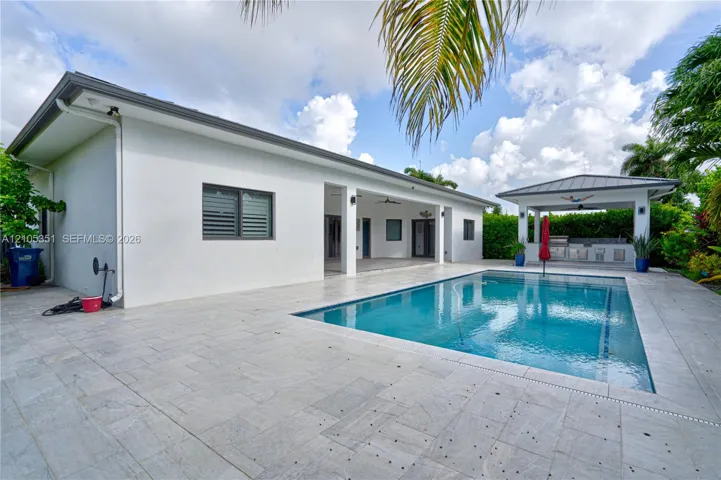 Backyard featuring a rectangular swimming pool, expansive stone-paver decking, covered patio, and a detached poolside pavilion with a built-in outdoor kitchen