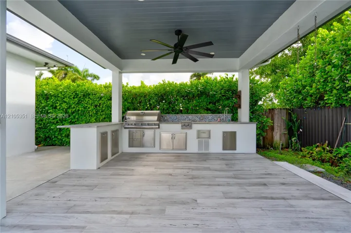 Covered outdoor kitchen featuring wood-finish tile flooring, built-in stainless steel grill, stone-style countertop, mosaic tile backsplash, and overhead ceiling fan