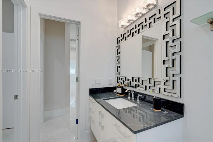 White vanity with dark stone countertop, undermount rectangular sink, chrome widespread faucet, geometric-patterned mirror, and four-light glass wall fixture