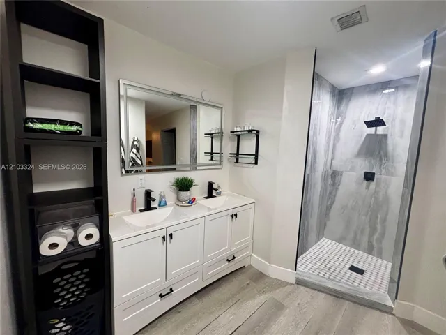 Dual-sink vanity with white cabinetry, rectangular vessel sinks, quartz countertops, and matte black fixtures, paired with a walk-in shower featuring large-format tile surround and wood-finish flooring