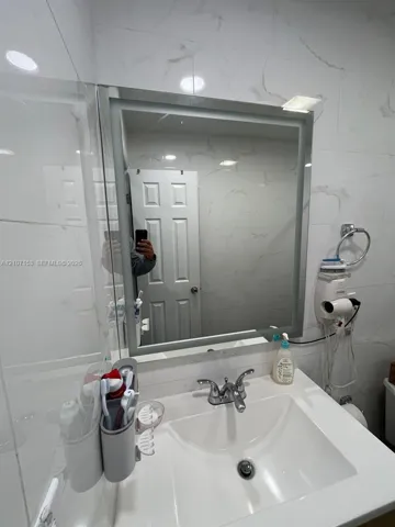 Illuminated rectangular wall mirror featuring large-format stone-patterned wall tile and a white vanity with an integrated basin and chrome dual-handle faucet