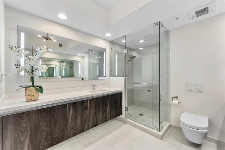 Floating vanity with wood-finish cabinetry, solid surface countertop, expansive wall-mounted mirror, glass-enclosed shower with tiled surround, and wall-mounted toilet