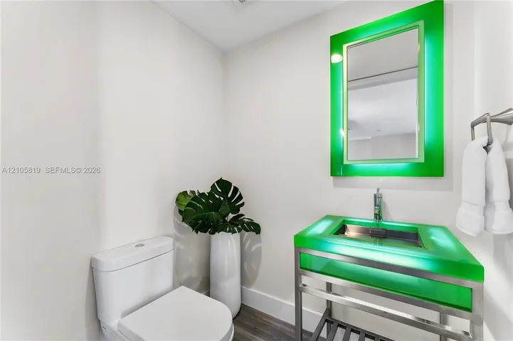 Floating vanity with an integrated green glass sink, polished metal frame, matching illuminated mirror, and wood-finish flooring