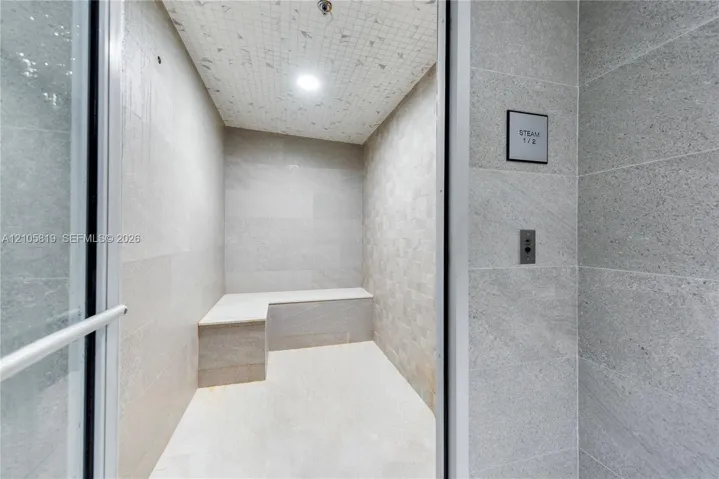 Steam room featuring large-format stone wall tile, mosaic ceiling tile, built-in stone bench, and glass-entry door with integrated control panel