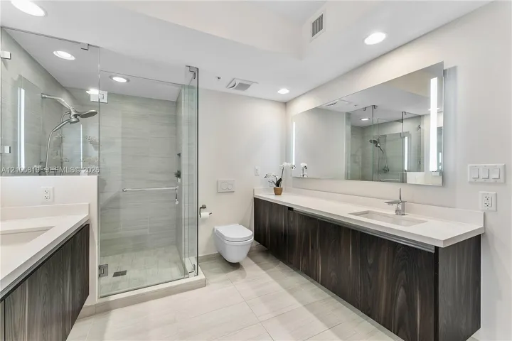 Floating dark wood-finish dual vanity with solid surface countertops, frameless glass-enclosed shower, wall-mounted toilet, and large integrated vanity mirror with LED lighting