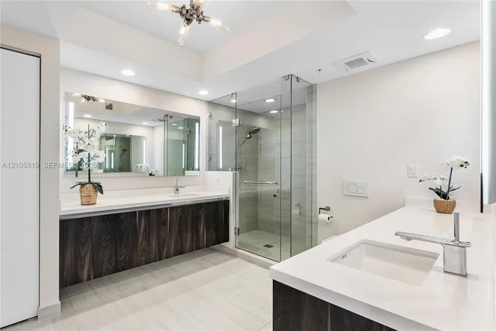 Dual floating vanities featuring dark wood-finish cabinetry, solid surface countertops, integrated rectangular sinks, and chrome fixtures, accompanied by a glass-enclosed walk-in shower with tile surround