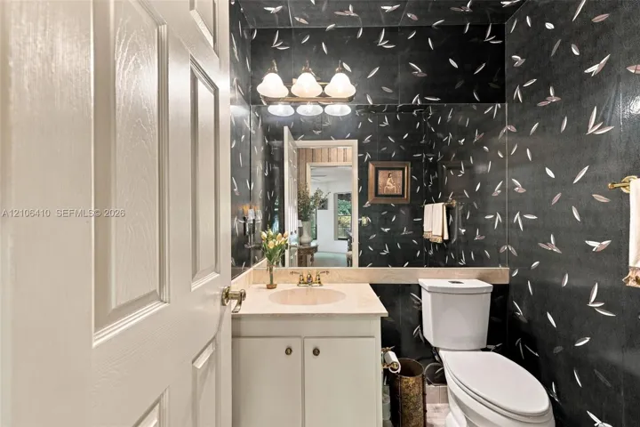 Powder room featuring patterned wall covering, stone-finish countertop with an integrated basin, vanity cabinet with dual-door storage, full-width vanity mirror, and three-light wall-mounted fixture