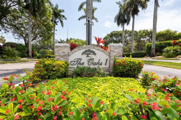 Community entrance sign featuring stacked stone columns, a curved metal plaque, and surrounding mature landscaping with flowering shrubs and palm trees