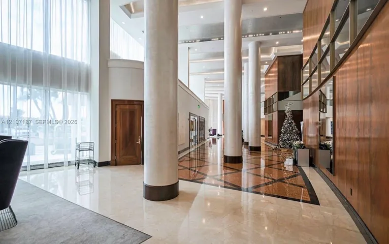 Grand lobby featuring floor-to-ceiling windows, soaring ceilings with recessed lighting, polished stone flooring with geometric tile inlay, full-height architectural columns, and wood-paneled wall accents