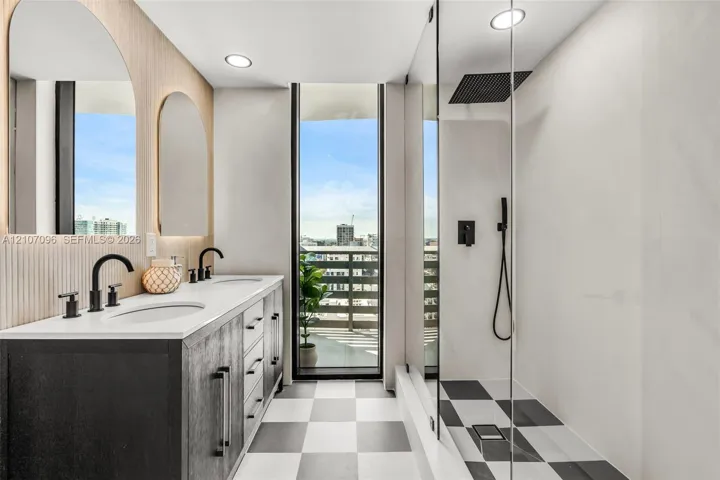 Double vanity with a white solid-surface countertop, dark cabinetry, arched wall mirrors, matte black fixtures, and a glass-enclosed shower featuring a rain-style showerhead and checkerboard tile flooring