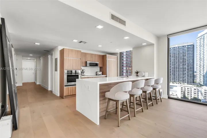 Modern kitchen featuring wood-finish cabinetry, a waterfall-edge quartz island, stainless steel built-in appliances, and floor-to-ceiling windows