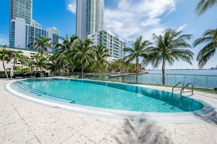 Oval swimming pool featuring a tiled finish, metal handrails, a light-colored aggregate pool deck, and a glass perimeter railing