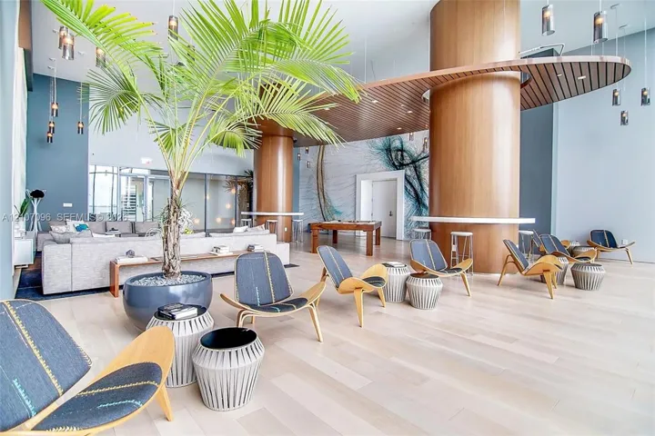 Open-concept common area featuring high ceilings, light wood-finish flooring, full-height wood-clad columns, a suspended circular wood-slat ceiling element, and decorative pendant lighting