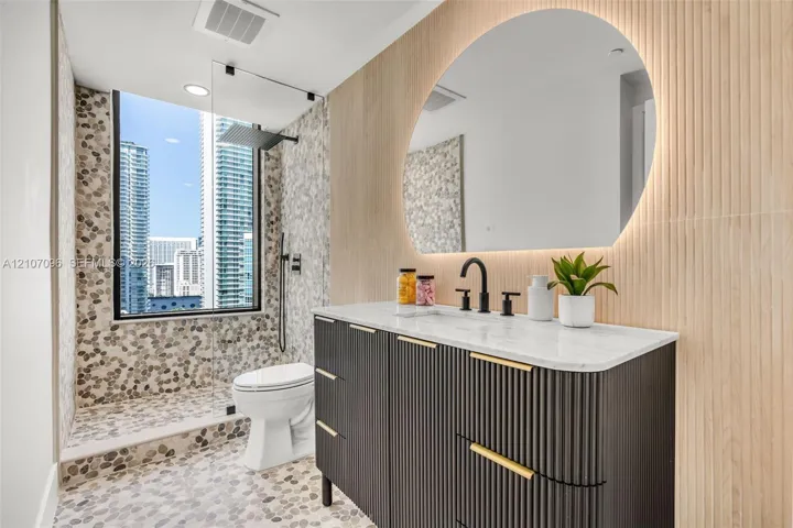 Custom dark fluted vanity with gold-tone hardware and light-toned stone countertop, integrated backlit arched mirror, floor-to-ceiling pebble tile shower surround, and matte black rain shower head